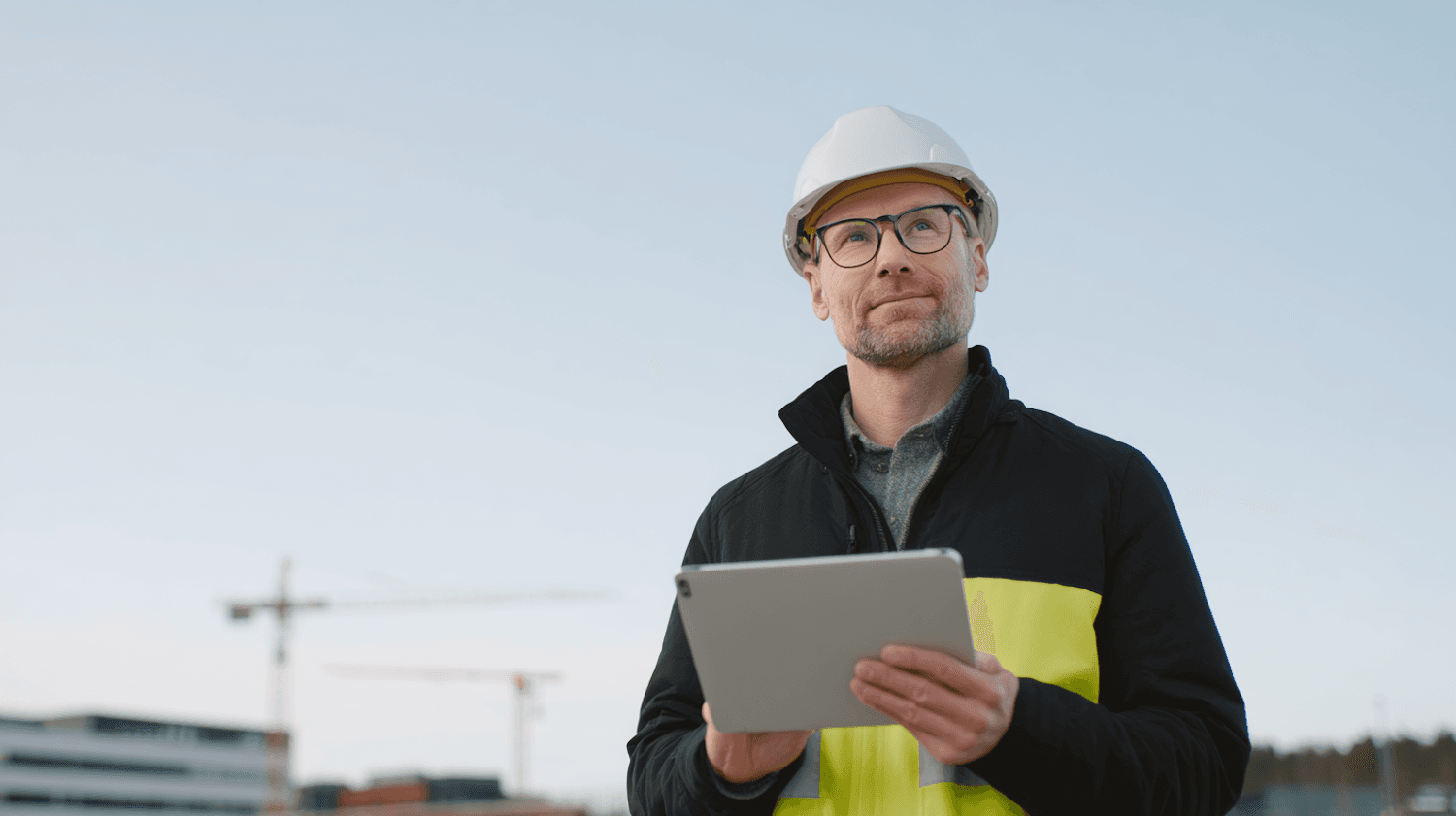 Site engineer in high-visibility gear holding a tablet with cranes behind